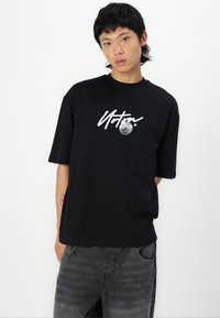 Black cotton t-shirt with short sleeves, featuring white graphic text and a moon design on the front. Pairing with grey denim pants.