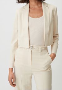 Beige kort jacka med smala slag, långa ärmar och en texturerad ribbad topp under. Bältesatta högmidjade byxor kompletterar setet.