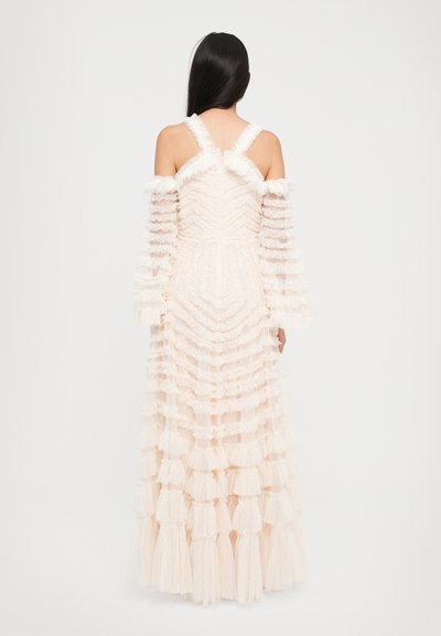 Vrouw met lang donker haar, gekleed in een witte jurk met ruches die tot op de vloer reikt en met off-shoulder mouwen, staande voor een effen witte achtergrond.