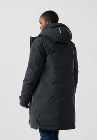 Persoon in een lange zwarte winterjas met capuchon en reflecterende details, die zijwaarts staat tegen een effen lichte achtergrond.