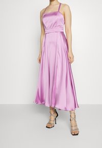Robe en satin sans manches, de couleur rose, avec un décolleté carré, une ceinture à la taille, et une jupe évasée avec des plis. Associée à des sandales à talons hauts avec des lanières.