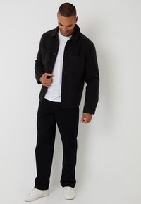 Mand iført sort jakke over hvid t-shirt, sorte bukser og hvide sneakers, stående mod en ensfarvet lysegrå baggrund.
