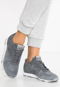 Personne portant des baskets Reebok Classic grises avec des semelles blanches et un pantalon de survêtement gris clair, les pieds positionnés avec l'un légèrement levé.