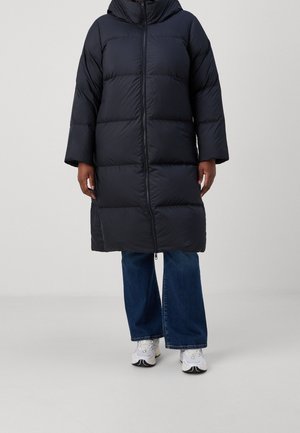 Eine Person trägt einen langen schwarzen Daunenmantel, weite blaue Jeans und weiße Sneakers und steht vor einem schlichten hellen Hintergrund.