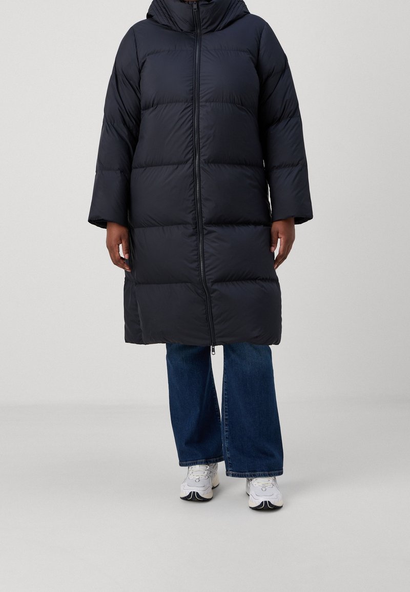 Persoon in een lange zwarte pufferjas, blauwe jeans met brede pijpen en witte sneakers, staand tegen een effen lichte achtergrond.