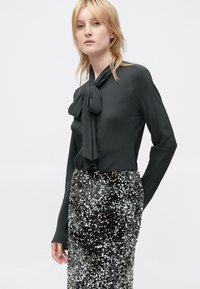 Blusa nera a maniche lunghe con un fiocco al collo, abbinata a una gonna a tubino in paillettes argento, caratterizzata da una silhouette aderente.