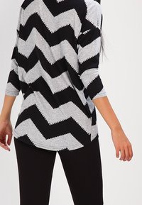 Svart och grå långärmad topp med zigzagmönster och vågiga kanter, kombinerad med svarta byxor, finns i bakvy.