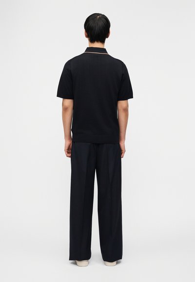 Άνδρας που στέκεται γυρισμένος προς τα πίσω, φορά μαύρο πουκάμισο με κοντά μανίκια, φαρδιά μαύρη παντελόνα και λευκά παπούτσια πάνω σε απλό ανοιχτό φόντο.