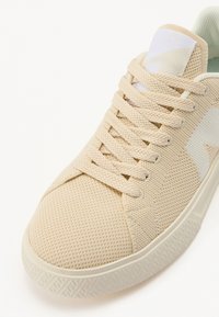 Sneaker in rete beige con punta rotonda, superficie testurizzata, lacci tono su tono e un logo bianco accentato sul lato. Suola in gomma con battistrada strutturato.