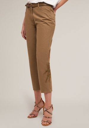 Pantalones cortos de color beige con una cintura ajustada y bolsillos laterales, que presentan un diseño de cuentas decorativas cerca del dobladillo. Combinados con sandalias de tacón alto y tiras.