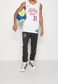 Jersey bianco senza maniche con testo rosso e blu, pantaloni neri con grafica di una rosa dorata e sneakers bianche con lettering audace. Tiene in mano un pallone da basket verde.