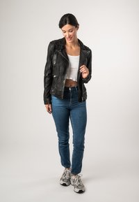 Schwarze Leder-Motorradjacke mit silbernen Reißverschlüssen, taillierte Passform, getragen über einem weißen Crop-Top, kombiniert mit blauen Jeans und Sneakers.