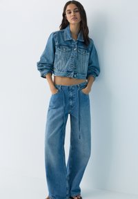 Femeie purtând o jachetă scurtă din denim albastru și blugi largi cu talie înaltă, de culoare albastru, stând cu mâinile în buzunare, sprijinită de un perete alb.