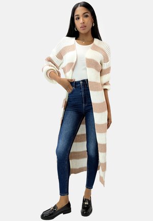 Lungo cardigan crema con strisce pesca, texture a costine, aperto davanti, indossato sopra una maglietta bianca aderente e jeans skinny scuri, abbinato a mocassini neri.