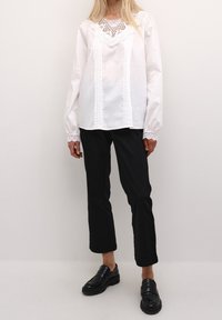 Bílá bavlněná blůza s krajkovým lemem, sklady a dlouhými rukávy, doplněná černými zkrácenými kalhotami a černými mokasíny.