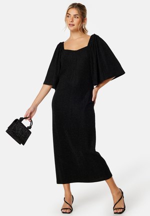Abito midi nero con scollatura quadrata, maniche corte a palloncino e tessuto strutturato. Gli accessori includono una piccola borsa nera e sandali con lacci.