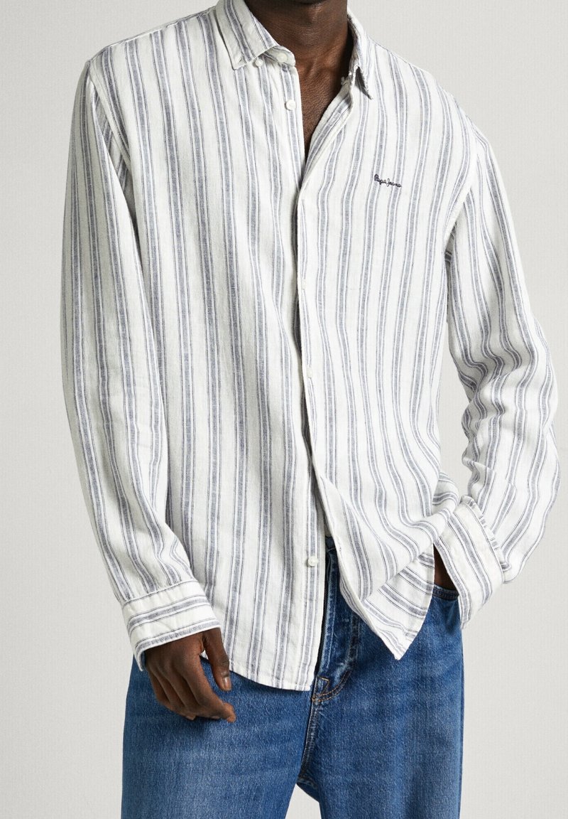 Homme portant une chemise blanche à boutons avec des rayures grises verticales et un jean bleu, main droite dans la poche, main gauche détendue sur le côté.
