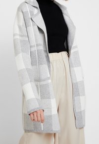 Frau trägt eine grau-weiße karierte Strickjacke, einen schwarzen Rollkragenpullover und beige hochgeschnittene Hosen vor einem schlichten Hintergrund.