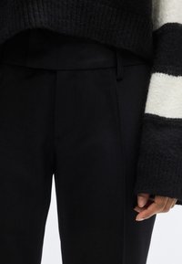 Μαύροι παντελόνες με κομψή σχεδίαση, κατασκευασμένοι από απαλό ύφασμα· συνδυασμένοι με χοντρό πουλόβερ πλεκτό σε μαύρο και άσπρο ριγέ σχέδιο.