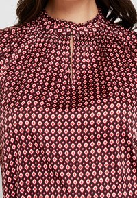 Hochgeschlossenes Blusentop mit einem geometrischen Rautenmuster in Rot, Weiß und Schwarz, ausgestattet mit einer Schlüsselöffnung und Raffung am Ausschnitt.