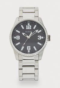 Montre en acier inoxydable argenté avec un cadran noir, de grands index horaires numériques et un bracelet en maillons argentés. Présente une lunette texturée.
