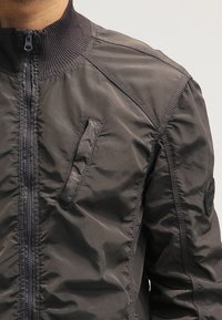 Mørkegrå bomberjakke lavet af letvægtsmateriale, med ribbet krave, lynlåsfront og en lille lynlås lomme på brystet.