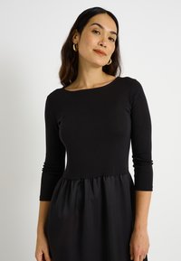 Vestido negro con parte superior acanalada y falda plisada de satén. Mangas tres cuartos, escote redondo y ajustado en la cintura.