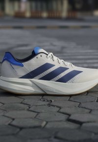 Chaussure de course blanche et bleue avec détail de semelle « LIGHTSTRIKE », sur un pavé gris, vue de côté.