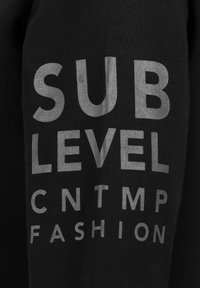 Zwarte stof met zilverbedrukte tekst: "SUB LEVEL CNTMP FASHION." De tekst heeft verschillende groottes en stijlen, wat zorgt voor een gedurfde contrast.