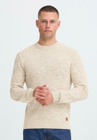 Man met kort haar en een oorbel, gekleed in een beige gebreide trui en blauwe jeans, staande tegen een effen witte achtergrond.