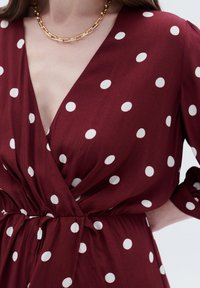 Burgunderfarbenes Wickelkleid mit weißen Polka-Dots, ausgestattet mit einem tiefen V-Ausschnitt, geraffter Taille und hochgekrempelten Ärmeln. Betont durch eine goldene Kettenhalskette.