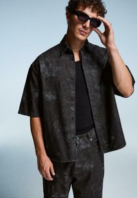 Uomo che indossa una camicia a maniche corte con motivo scuro e pantaloni, canottiera nera, occhiali da sole e che tocca gli occhiali da sole con la mano su uno sfondo neutro.