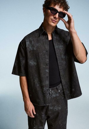 Uomo che indossa una camicia a maniche corte con motivo scuro e pantaloni, canottiera nera, occhiali da sole e che tocca gli occhiali da sole con la mano su uno sfondo neutro.