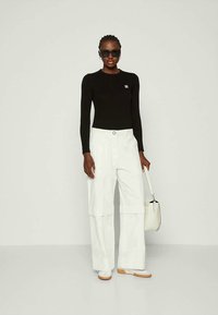 Mulher vestindo uma blusa preta de manga longa, calças brancas largas, sapatos brancos, óculos de sol, e segurando uma bolsa branca contra um fundo simples.