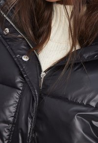 Schwarze Pufferjacke mit hohem Kragen, silbernem Reißverschluss und Druckknöpfen. Hergestellt aus glänzendem, glattem Material mit gestepptem Muster.