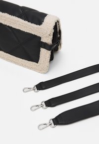 Svart väska med en ullik beige kant, i en kviltad design. Tre avtagbara svarta remmar med silverdetaljer ligger bredvid.