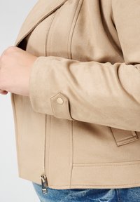 Beige jas van nep suède met lange mouwen, ritssluiting en drukknoopmanchet. Heeft een gladde textuur en een gestructureerd ontwerp.