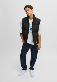 Black padded vest with a high collar, worn over a white t-shirt featuring a graphic design. Paired with dark denim jeans and white sneakers.