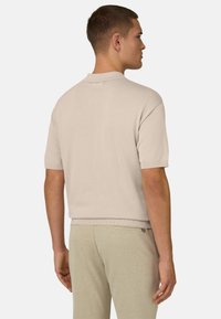 Homme portant un polo en maille beige à manches courtes et un pantalon beige assorti, debout de dos avec la tête légèrement tournée vers la droite.