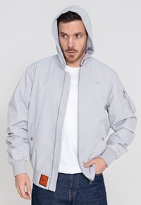 Graue Bomberjacke mit einem gesteppten Design, grauer Kapuze im Sweatshirt-Stil, seitlichen Taschen und einem orangen Etikettakzent. Mit elastischen Bündchen und Saum.