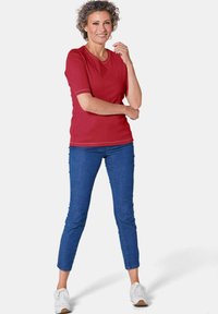 Rotes Kurzarm-Oberteil mit rundem Hals, kombiniert mit eng anliegenden blauen Jeans und weißen Turnschuhen. Das Oberteil hat einen dezenten kontrastierenden Saum.