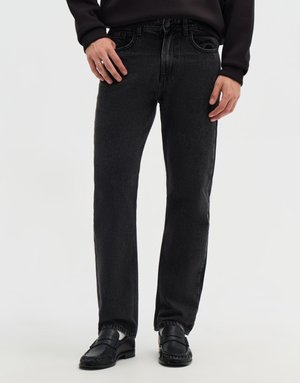 Uomo che indossa jeans neri a gamba dritta, mocassini neri, calzini bianchi e una camicia nera a maniche lunghe, in piedi davanti a uno sfondo uniforme.
