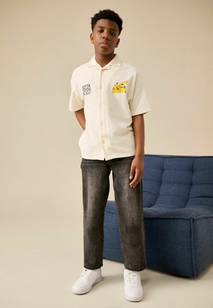 Dreng står ved en blå polstret stol iført en cremefarvet skjorte med Pikachu og teksten "Gotta Catch ’Em All", sorte jeans og hvide sneakers.