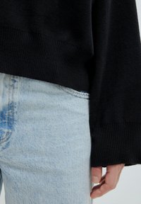 Schwarzer gerippter Pullover mit lockerem Schnitt, kombiniert mit hellblauen, zerstörten Jeans. Nahaufnahme einer Hand auf den Jeans, die den Stoff und die Textur zeigt.