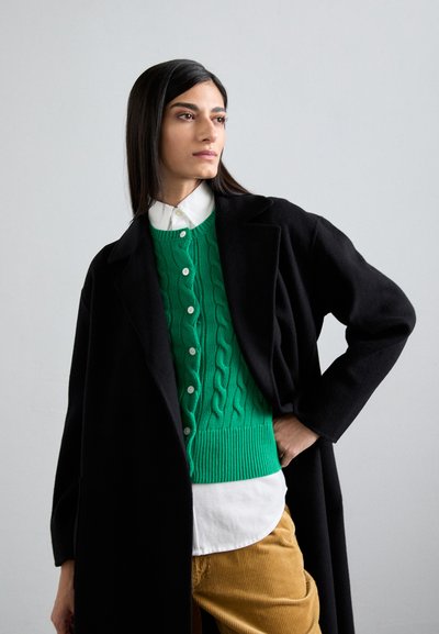 Groene gebreide trui met kabelpatronen, zwarte jas en daaronder een witte shirt. Model draagt bruine corduroy broek. Gladde, matte texturen.