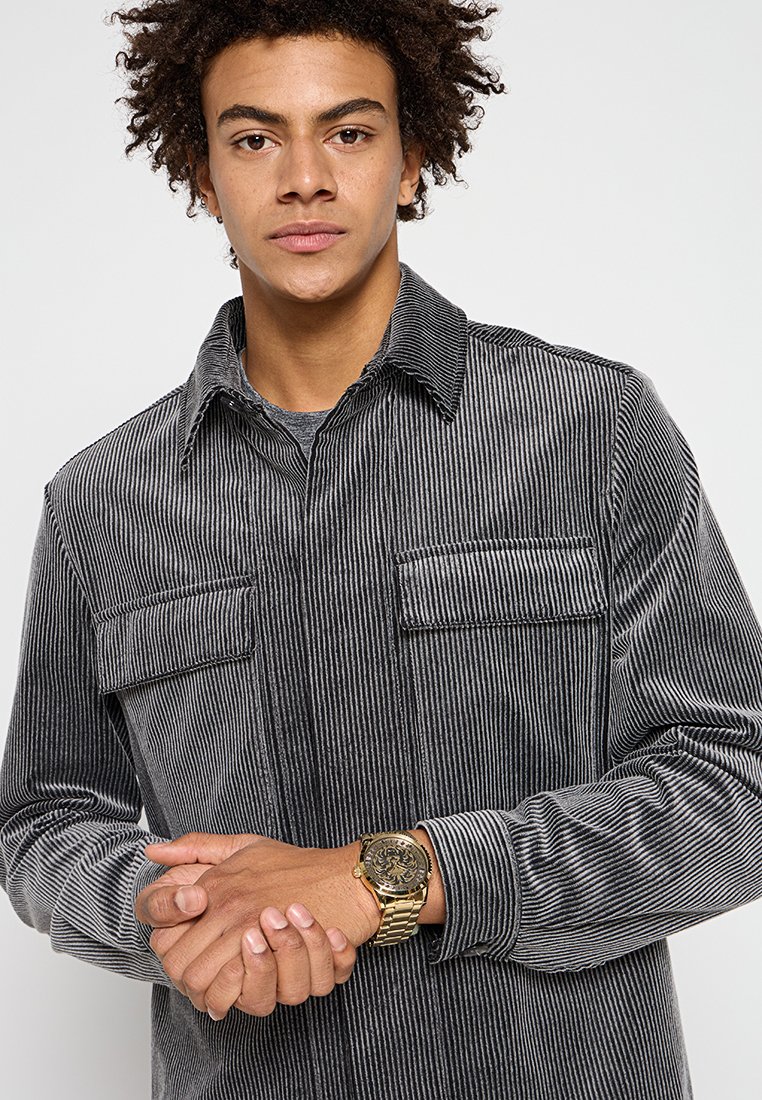 Joven vistiendo una camisa de pana gris con rayas y un reloj de pulsera dorado, juntando las manos frente a un fondo blanco liso.