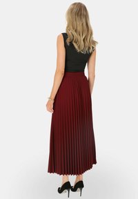 Burgundowa, plisowana spódnica maxi z wysokim stanem, zestawiona z czarną, bezrękawnikową bluzką. Modelka nosi czarne szpilki.