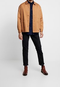 Vaaleanruskea kangastakki, jossa on kaulus, napitus ja kaksi etutaskua, päällä tummansininen paita sekä tummat farkut ja ruskeat saappaat.