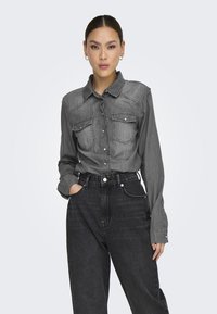 Camisa de mezclilla gris con mangas largas, cuello y dos bolsillos frontales, combinada con pantalones vaqueros negros de cintura alta. Textura suave y diseño estructurado.