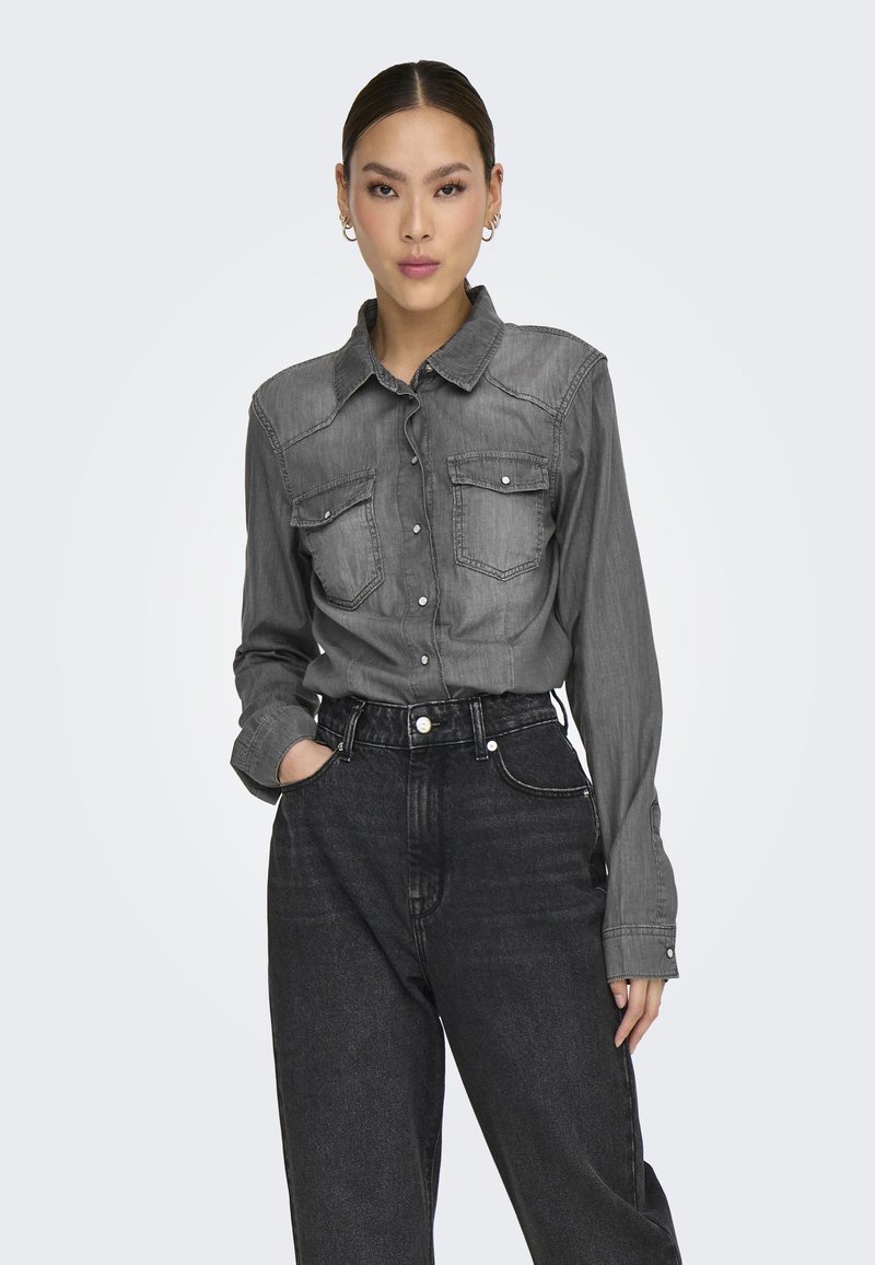 Camisa de mezclilla gris con mangas largas, cuello y dos bolsillos frontales, combinada con pantalones vaqueros negros de cintura alta. Textura suave y diseño estructurado.